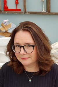 Artist Sam Larter. The image shows artist Sam Larter with medium length dark hair and black framed glasses with a dark top and necklace sat in her studio in front of her workbench.